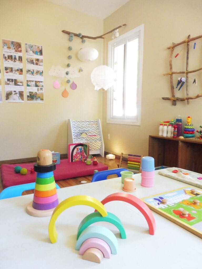 A colorful children's playroom in Honduras features a pink mat on the floor with toys, including stacking rings, a wooden abacus, and a board game, alongside decorated walls with photos, paper clouds, and hanging lanterns, set near a window that lets in natural light.