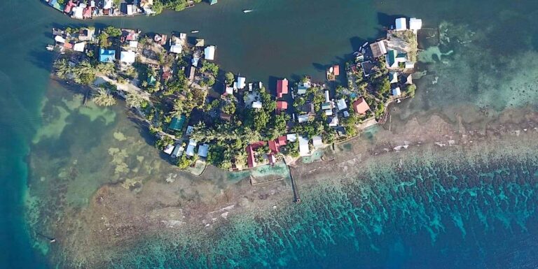 An aerial view of a small island in Roatan, Honduras, showing clusters of houses surrounded by lush greenery, water on all sides, and coral reefs along the shoreline.