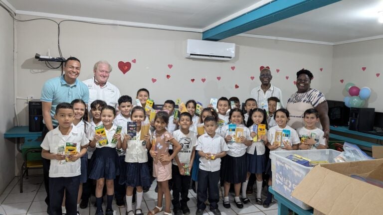 A group of smiling children and adults gather indoors in Roatan, Honduras, holding school supplies such as pencils and notebooks, with heart-shaped decorations on the wall and balloons in the background.