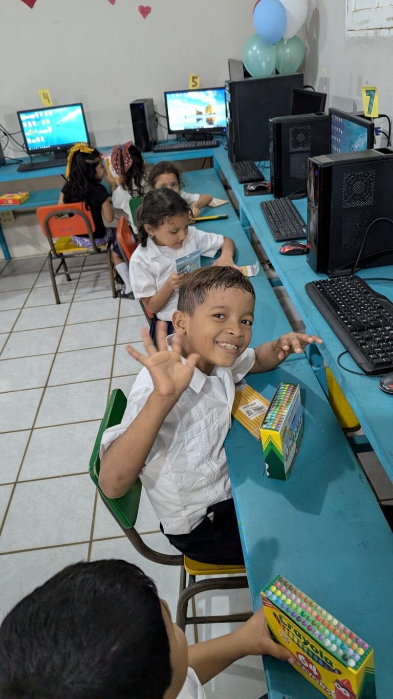Children sitting at a turquoise table with desktop computers and monitors, participating in a computer activity or game, with balloons and colorful decor indicating a festive setting in Roatan, Honduras.