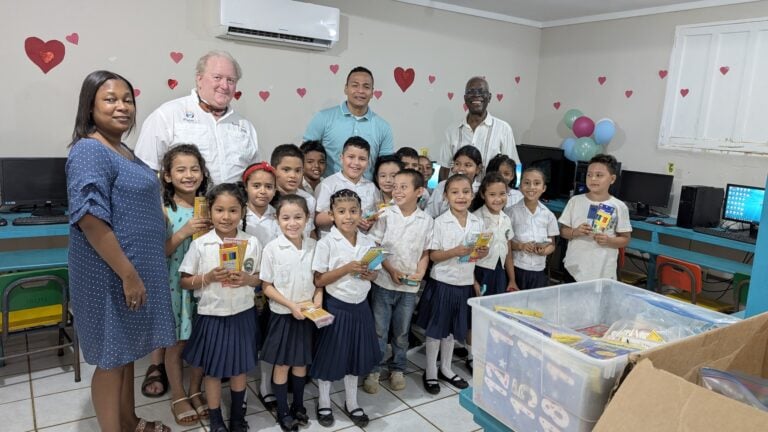 A group of children and adults gather in a classroom on Roatan, Honduras, holding school supplies with computer stations, tablets, and colorful balloons in the background.