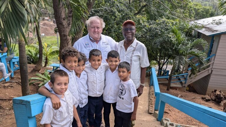 A group of children and two adult men, one wearing a white shirt with "Roatan" branding, pose together outdoors among lush greenery and blue stairs in Roatan, Honduras.