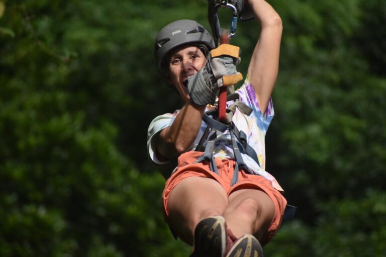 A woman wearing a helmet and gloves is zip-lining through a lush green forest in Roatan, Honduras.