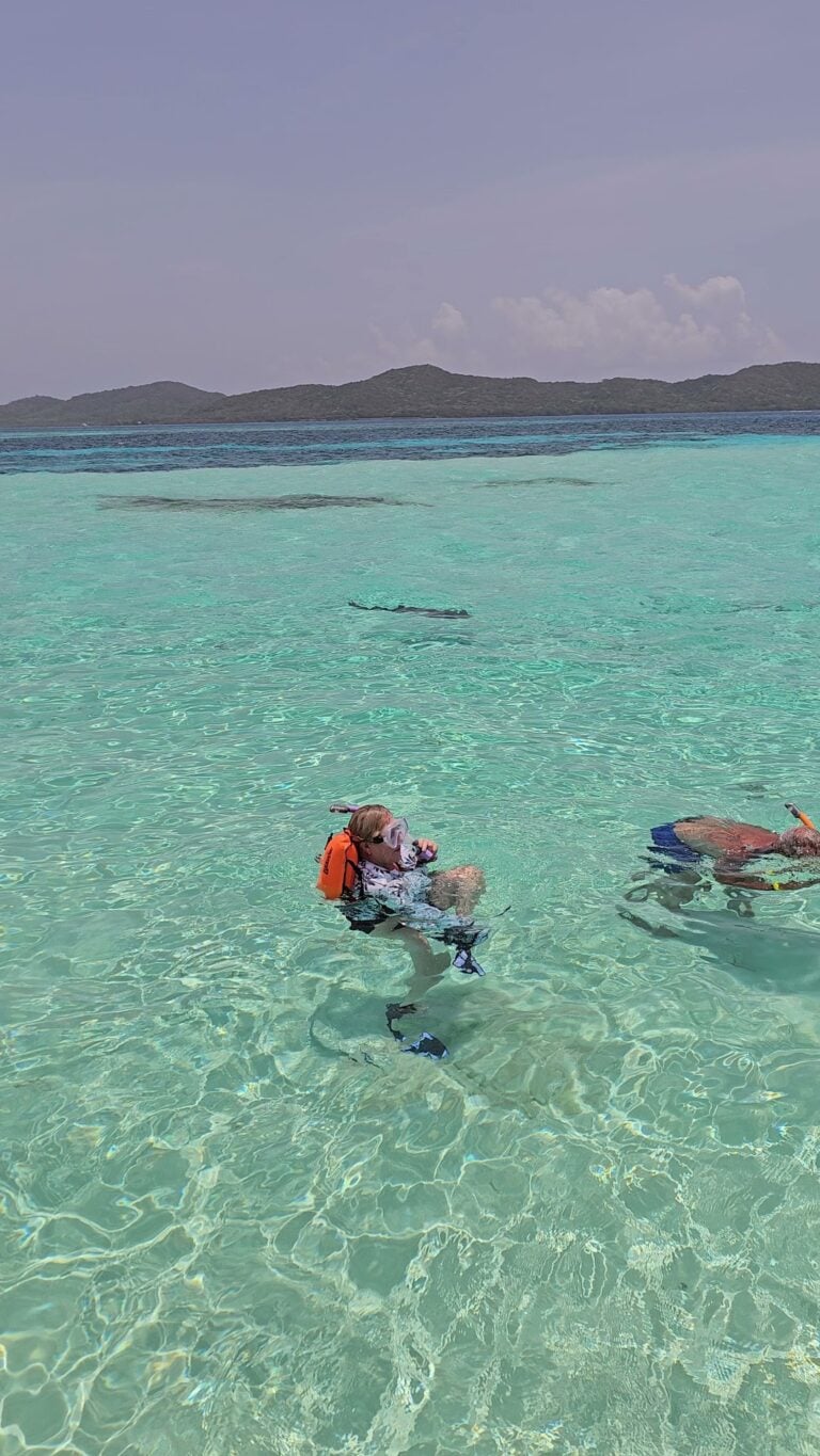 Two people are snorkeling in the clear turquoise waters of Roatan, Honduras, with lush green islands visible in the background.