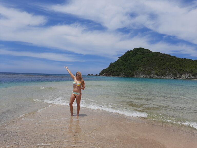 A woman in a colorful bikini holding a drink stands at the edge of the clear, gentle waters of Roatan, Honduras, with lush green island hills and a partly cloudy sky in the background.