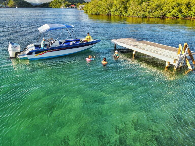A group of people swimming and relaxing in clear green waters near a wooden dock in Roatan, Honduras, with a small motorboat anchored nearby and lush green trees along the shoreline.