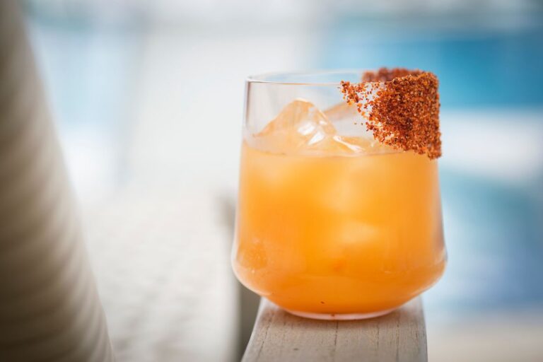 A tropical cocktail with an orange hue and ice cubes is garnished with a chili rim and sits on a wooden surface, set against a blurred background of Roatan, Honduras.