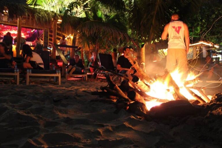 People gather around a bonfire on the sandy beach at night in Roatan, Honduras, with a man standing near the fire and others sitting on benches under palm trees at a lively beachside bar or restaurant.