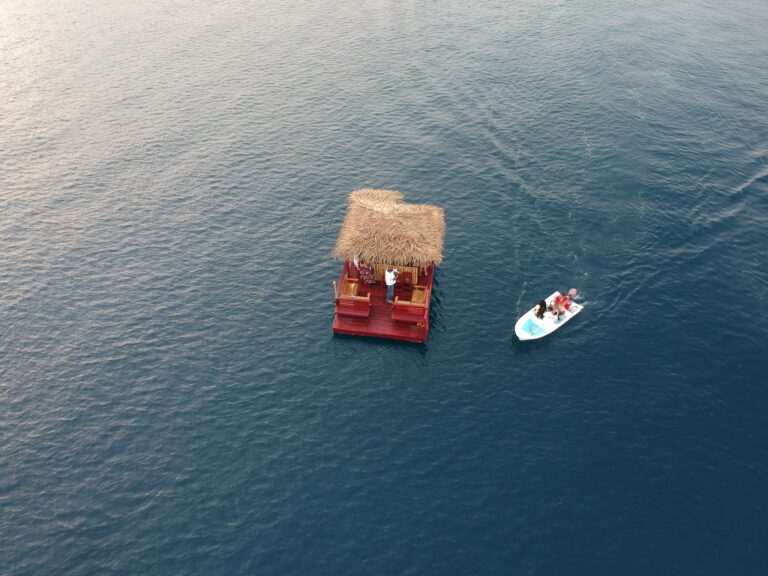 A small thatched-roof boat with two people on a platform and another boat with four passengers in life jackets on the clear waters of Roatan, Honduras.