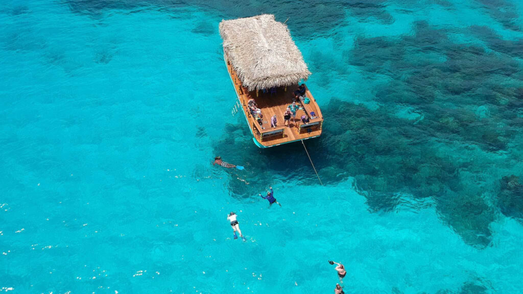 A group of people on a thatched-roof boat enjoying a scenic cruise over clear turquoise waters off Roatan, Honduras.