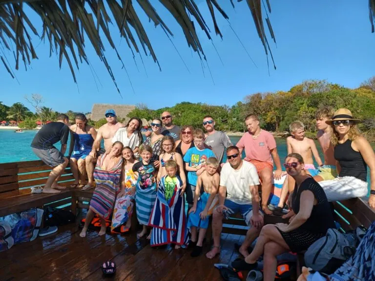A large group of people, including children and adults, gather on a wooden boat deck with a thatched roof in Roatan, Honduras, enjoying sunny weather and surrounded by lush green trees and calm water.
