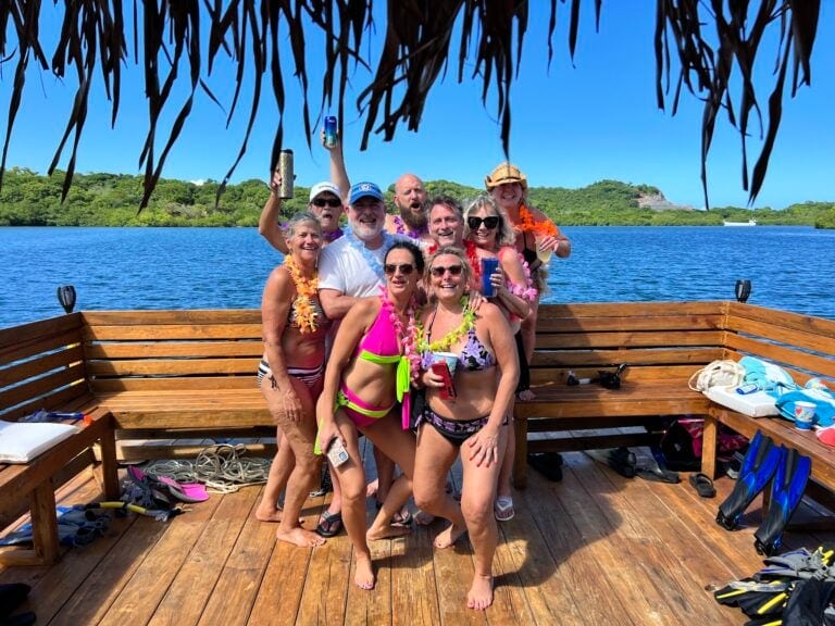 A group of friends wearing colorful swimsuits and leis pose on a wooden boat deck with tropical scenery, trees, and a clear blue sky in Roatan, Honduras.