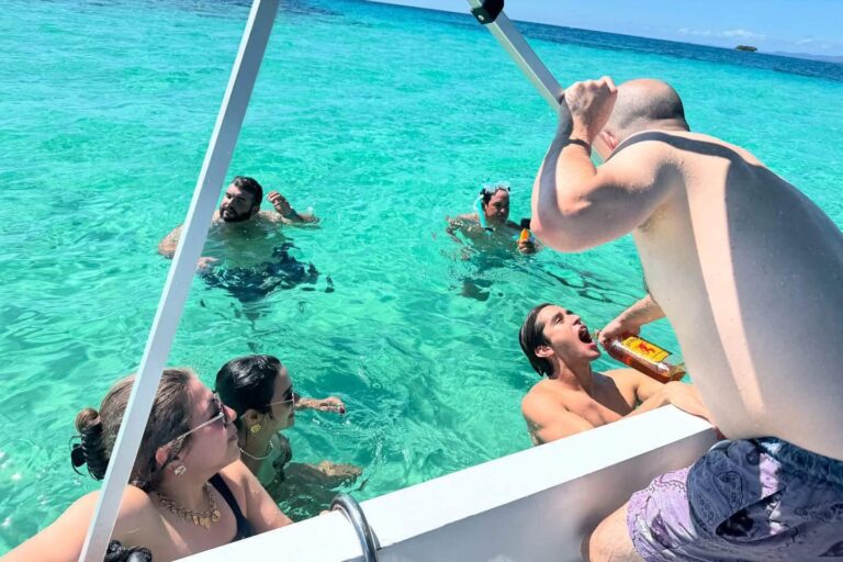 A group of people enjoying drinks and socializing in the clear turquoise waters near a boat in Roatan, Honduras, with an open sky and distant landmass in the background.