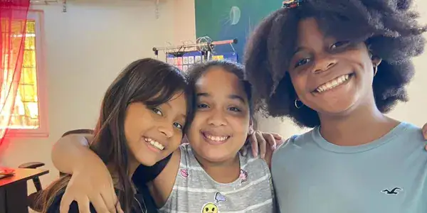 Three smiling young girls with dark hair and bright expressions pose closely together inside a colorful room in Roatan, Honduras.