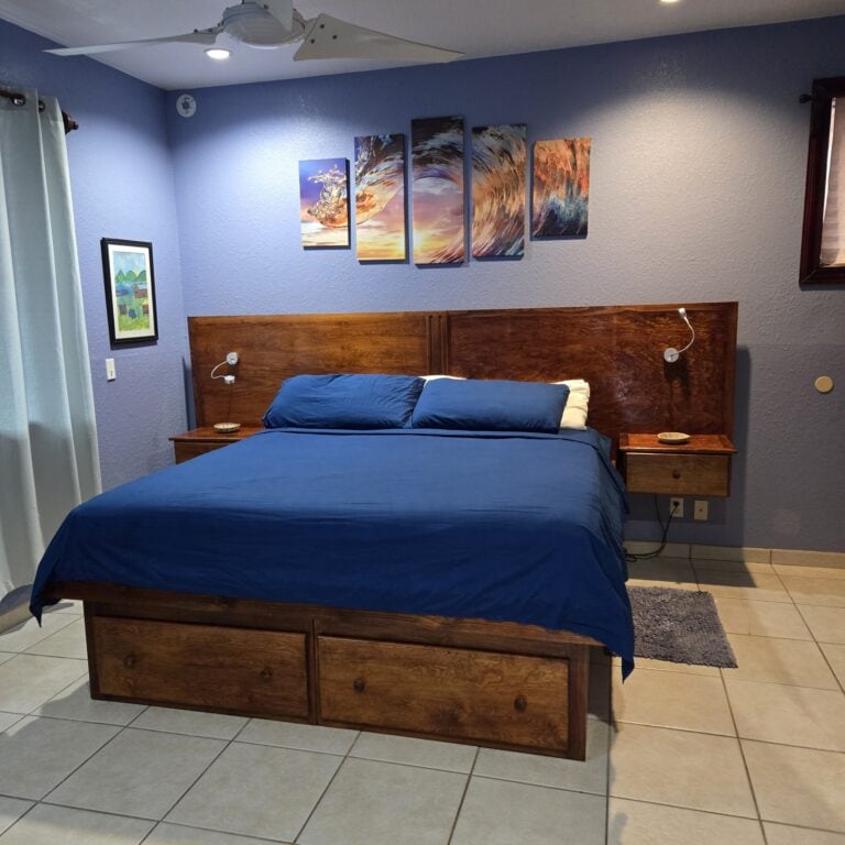 Bedroom with a wooden bed frame, blue bedding, and artwork depicting ocean waves on the blue wall, located in a Roatan, Honduras villa or hotel.