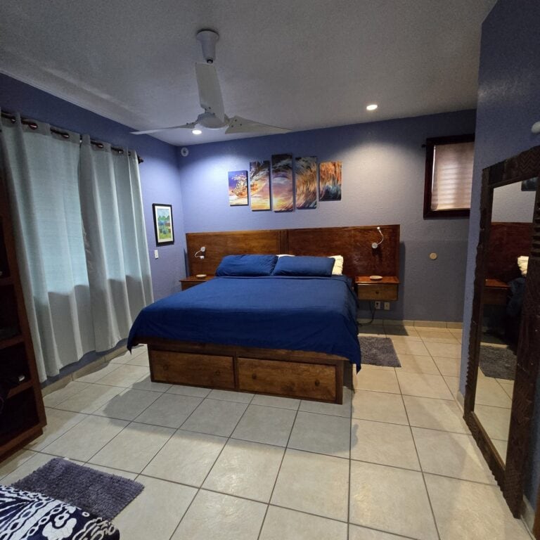 A cozy bedroom with a wooden bed frame, blue bedding, and sea-themed artwork on the blue painted wall in a Roatan, Honduras vacation rental.