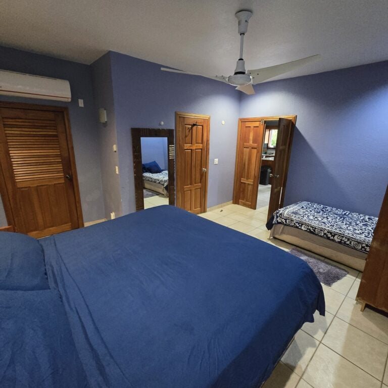 Interior view of a bedroom in Roatan, Honduras, featuring two beds with blue bedding, wooden doors, a full-length mirror, and a ceiling fan with an air conditioning unit on the wall.