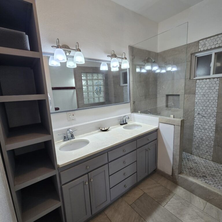 Dual bathroom sinks with a large mirror, overhead lights, and a glass shower enclosure with beige tile and small hexagonal floor tiles in Roatan, Honduras.