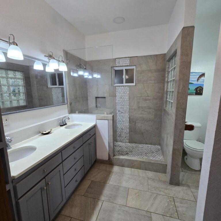 Modern bathroom with dual sinks, large mirror, glass block window, and walk-in shower with beige tiles on Roatan Honduras.