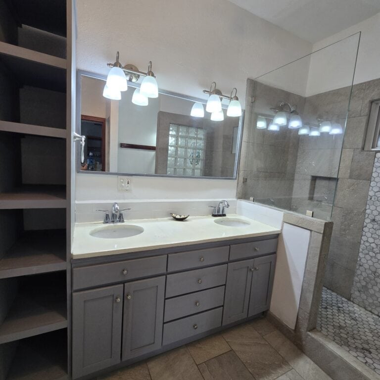 A modern bathroom in Honduras featuring dual sinks with a large mirror and contemporary light fixtures, adjacent to a glass-enclosed shower with beige tile walls.