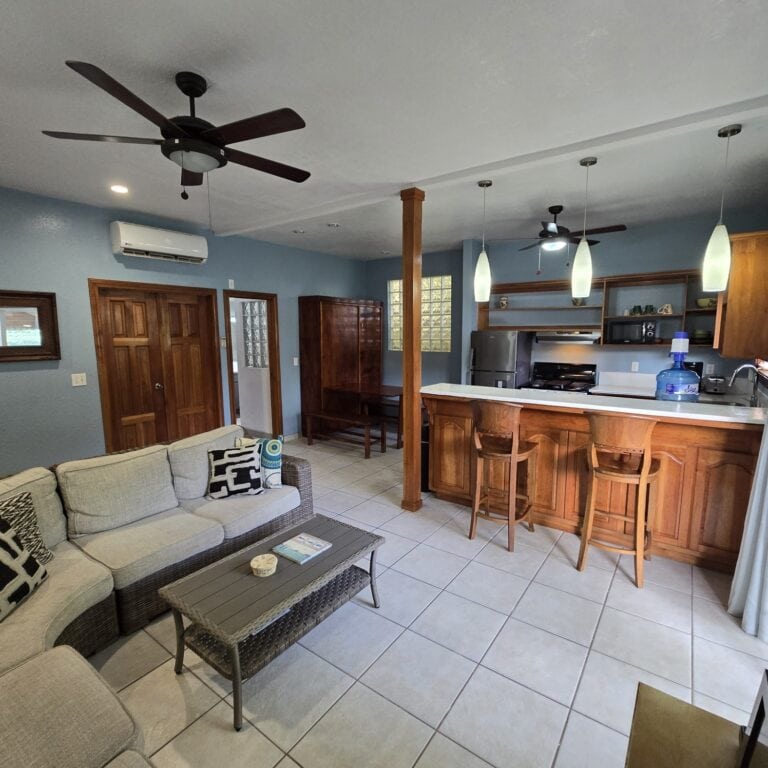 Interior view of a cozy living room in a Roatan vacation home with a kitchen area in Honduras featuring wooden cabinets, hanging pendant lights, ceiling fans, and a sofa with decorative pillows.