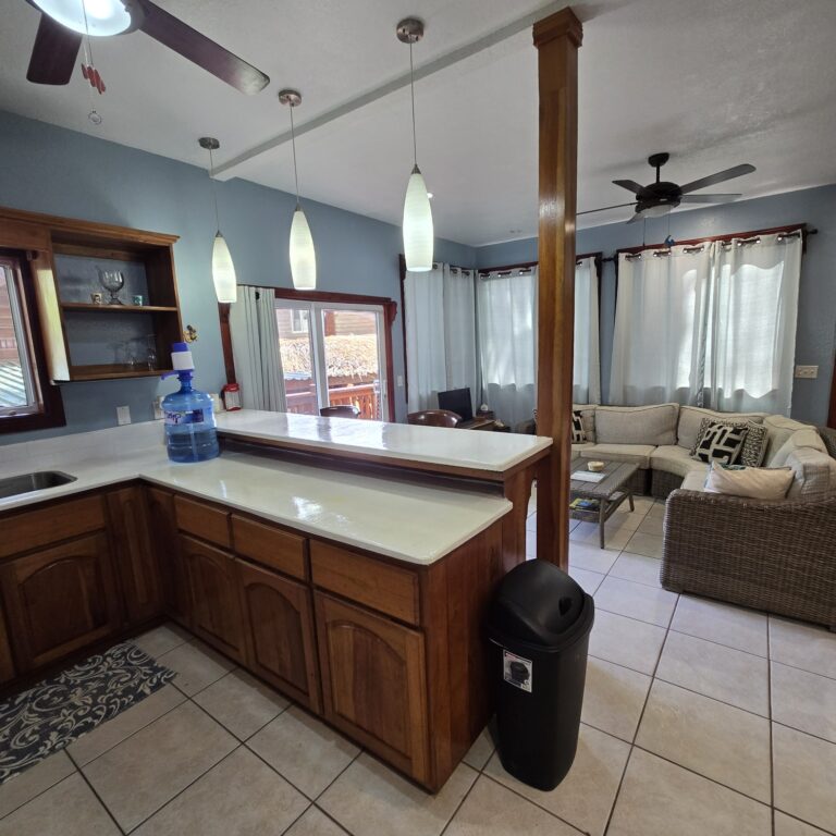 Open-concept living room and kitchen in a house on Roatan, Honduras, with a kitchen island, wicker sofa, and large windows with white curtains.