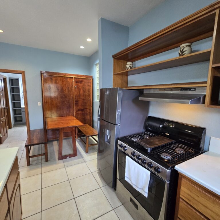 Inside view of a kitchen with blue walls, wooden cabinets, a stainless steel refrigerator, a gas stove, and open shelving with decorative pottery in Roatan, Honduras.