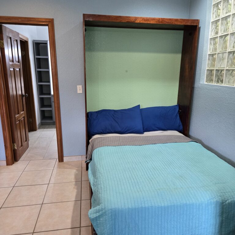 A cozy bedroom featuring a neatly made bed with blue pillows and a light blue quilt, set against a green accent wall in a guesthouse on Roatan, Bay Islands, Honduras, with a window made of glass blocks providing natural light.