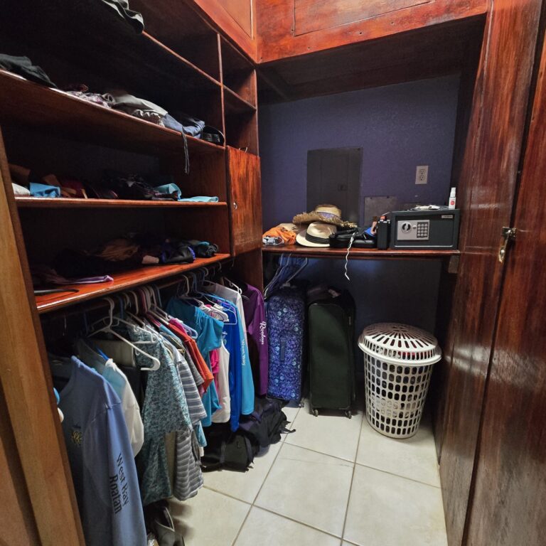 A compact closet with wooden shelves, hanging clothes, and travel suitcases inside a property on Roatan, Honduras.