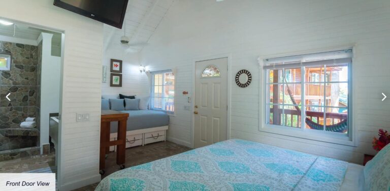 Interior of a cozy bedroom in Roatan, Honduras, featuring a white wooden wall, a large window with a view of a balcony and lush greenery, a bed with patterned bedspread, and decorative wall art with a tropical theme.