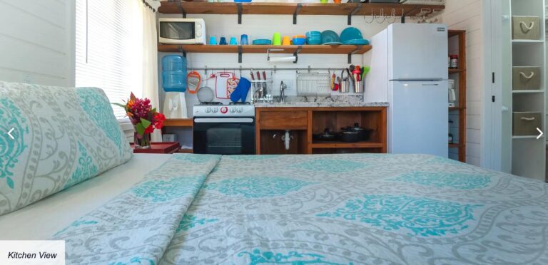 Cozy kitchen in a Caribbean-style home on Roatan, Bay Islands, Honduras featuring white wooden walls, open wooden shelves with colorful dishes, a small stove, a microwave, and a refrigerator.