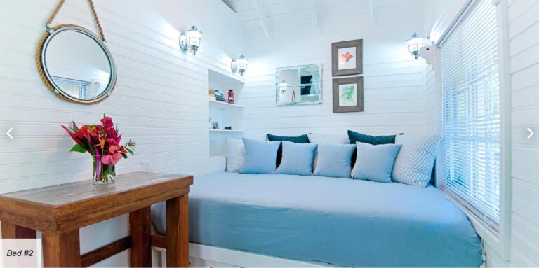 A cozy white-walled living room on Roatan with a large light blue sofa adorned with multiple blue and gray cushions, decorated with tropical-themed framed artwork and a mirror, illuminated by wall-mounted lantern-style lights, and featuring white blinds on the windows showing sunlight filtering through.