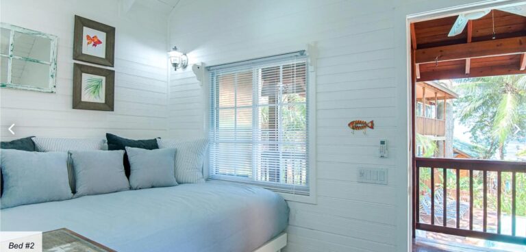 A cozy bedroom in a Roatan vacation home featuring white wooden walls, a large bed with multiple pillows, and tropical artwork on the wall, with a view of the balcony and lush greenery outside in Honduras.