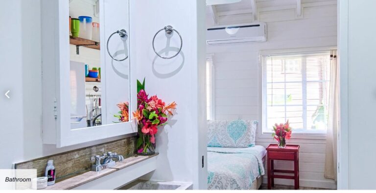 A cozy bedroom in Roatan, Honduras featuring white wooden walls, a bed with a patterned bedspread, and large windows with blinds allowing natural light to fill the room, decorated with colorful flowers on a small red table.