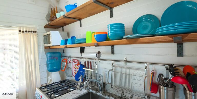 Open kitchen in Roatan, Honduras featuring white paneled walls, wooden shelves with colorful dishes and a microwave, next to a window with white curtains.