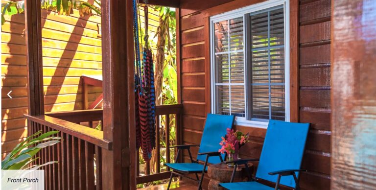 A wooden front porch in Roatan, Honduras, featuring blue patio chairs and a small table with a colorful flower arrangement, surrounded by tropical plants and banana trees.