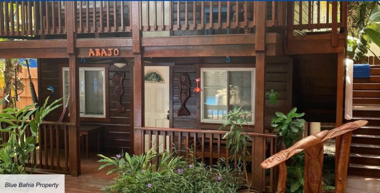 A wooden two-story building with a balcony and tropical decor located in Roatan, Honduras, featuring carved fish decorations on the exterior wall and lush green plants nearby.