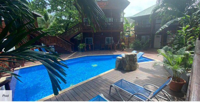 A rectangular outdoor swimming pool surrounded by a wooden deck and lush tropical plants in Roatan, Honduras, with wooden cabins and green trees in the background.