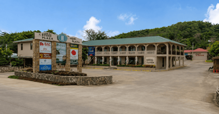The Coral Stone Plaza in Roatan, Honduras, features a two-story building with a green metal roof and arched verandas, surrounded by lush green hills and a clear blue sky.