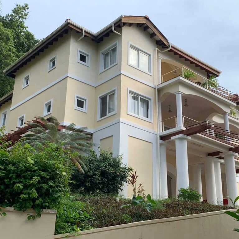 A large cream-colored multi-story villa with arched balconies, white columns, and tropical landscaping in Roatan, Honduras.