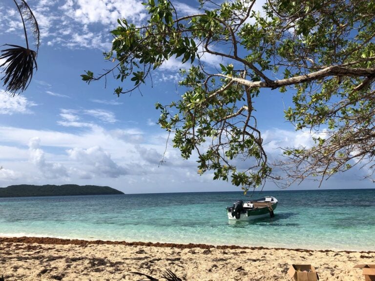 A small boat anchored near the sandy beach at Roatan, Honduras, with lush green trees and a partly cloudy sky in the background.