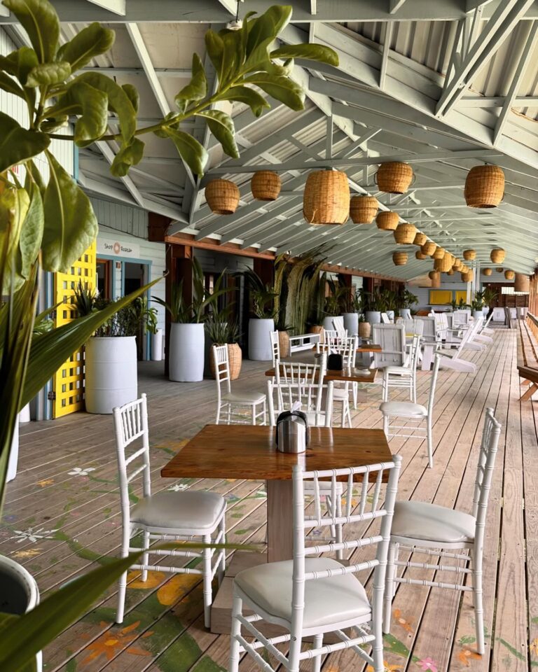 Open-air dining area with white chairs and wooden tables decorated with plants and hanging wicker lamps in Roatan, Honduras.