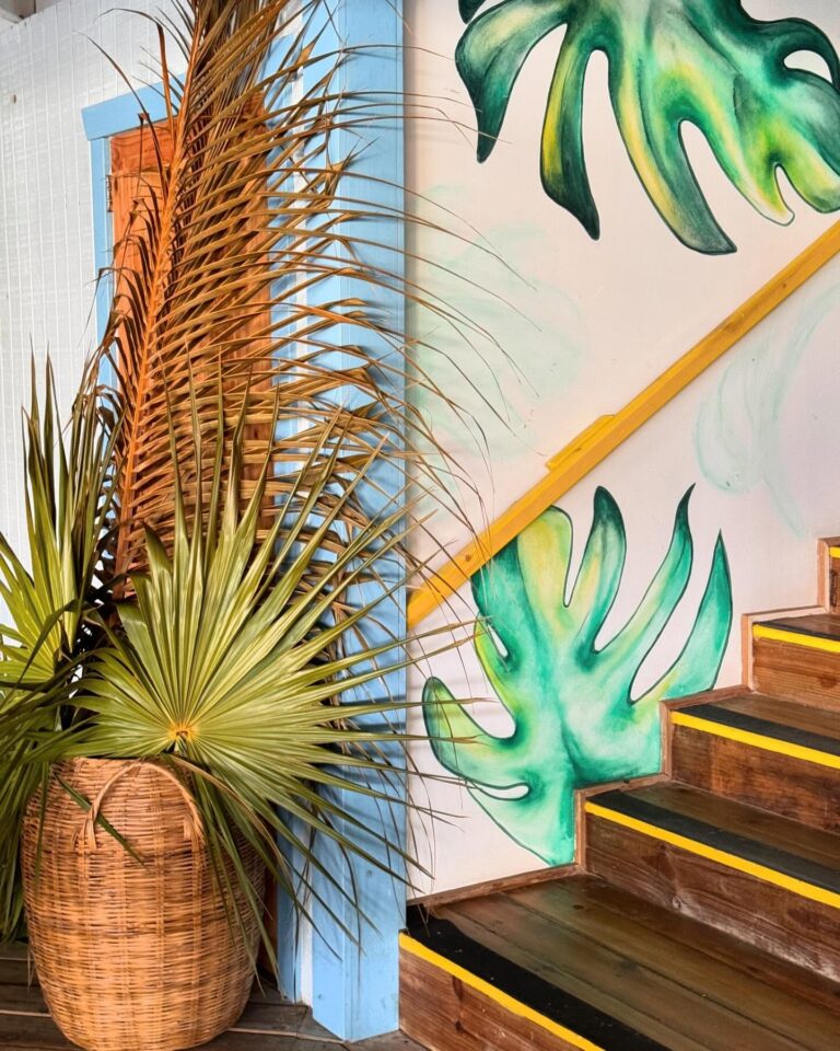 Colorful tropical-themed mural with large green Monstera leaves painted on a white wall, a staircase with yellow and black treads, and lush potted palm plants in woven baskets on Roatan, Honduras.