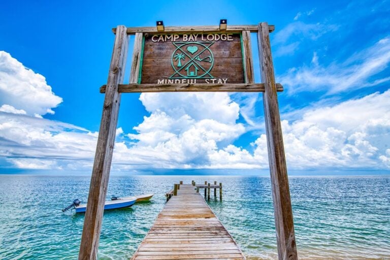 A wooden pier extends out over clear turquoise waters with a sign reading 'Camp Bay Lodge' and 'Mindful Stay' above the entrance, against a backdrop of blue sky and scattered white clouds in Roatan, Honduras.