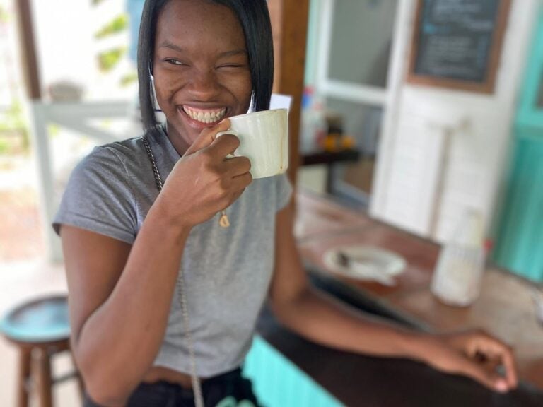 A woman with a broad smile holds a white ceramic mug while standing inside a cozy cafe in Roatan, Honduras, with colorful wooden decor and a chalkboard menu in the background.