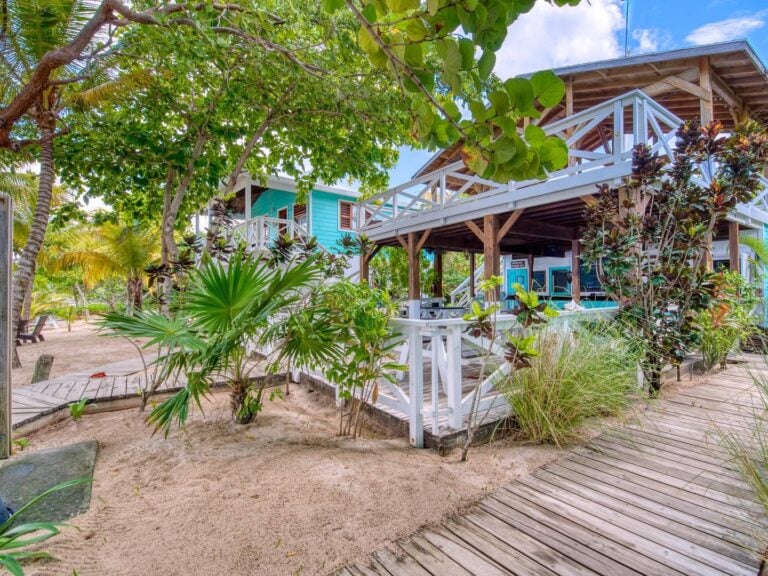 Colorful wooden beach houses with white railings surrounded by lush tropical plants and trees on Roatan in Honduras, with sandy pathways and a bright blue sky in the background.