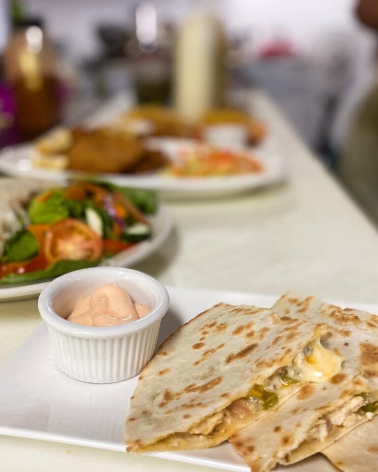 Close-up of a meal featuring a grilled quesadilla with cheese and jalapenos, alongside a fresh garden salad with tomatoes, lettuce, and carrots, and a small dish of pink sauce, set on a white plate in a restaurant on Roatan, Honduras.
