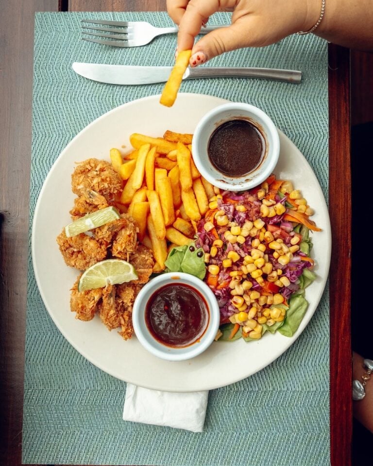 A plate of fried chicken tenders with lemon wedges, French fries, a fresh vegetable salad with corn, and two small bowls of dipping sauces, all set on a green placemat at a dining table in Roatan, Honduras.