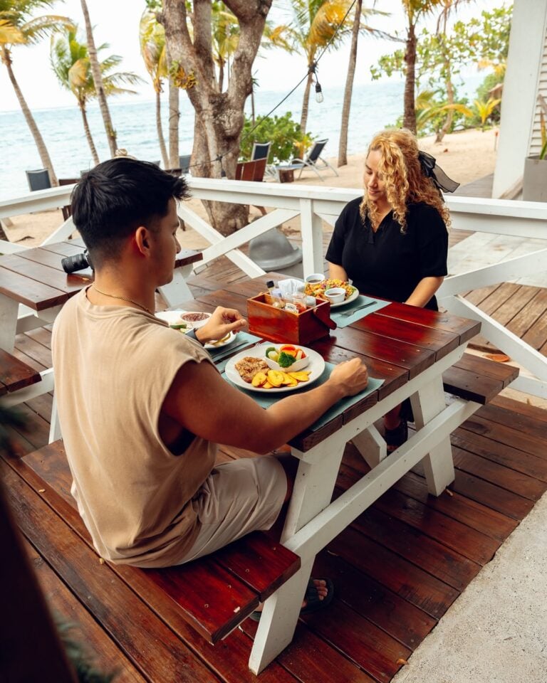 A man and a woman enjoying a meal at an outdoor restaurant on Roatan's beachside with palm trees and the sea in the background in Honduras.