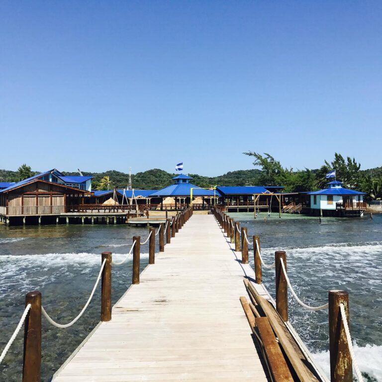 Wooden pier leading to a waterfront resort with blue-roofed bungalows and hammocks on Roatan, Honduras.